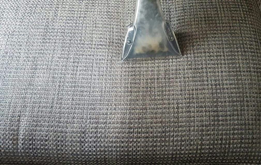 Close-up of a professional upholstery cleaning tool lifting dirt from grey woven fabric